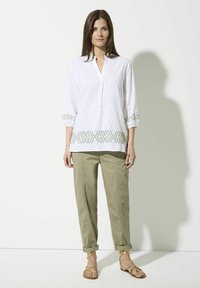 Weißes Leinenhemd mit grünem besticktem Muster am Saum und an den Ärmeln, kombiniert mit khakifarbenen Hosen und goldenen Sandalen.