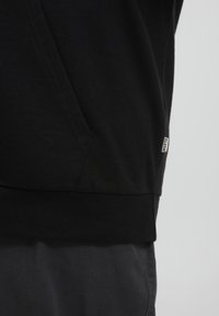 Sudadera de algodón negra con un bolsillo tipo canguro, dobladillo y puños acanalados, con una pequeña etiqueta de marca en el lado izquierdo. Textura suave.