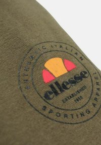 Logo de la marque Ellesse sur tissu vert olive, avec un design de demi-balle de tennis en orange et rouge accompagné d'un texte circulaire noir.
