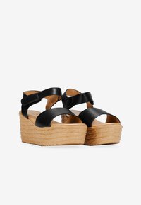 Sandales compensées noires avec des lanières, dotées d'une semelle en tan texturée. Elles comportent des lanières synthétiques lisses et un design ouvert à l'avant.