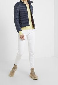 Doudoune bleue marine avec une fermeture éclair à l'avant et un col montant, portée sur un pull jaune clair, associée à un pantalon blanc et des bottes beige.