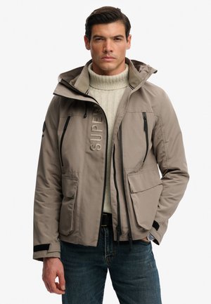 Mand iført beige hættetrøje med flere lynlåslommer over en cremefarvet kabelstrikket rullekravetrøje og blå jeans.
