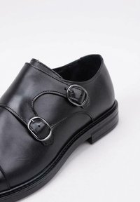 Zapato de cuero negro con dos correas de hebilla plateada, punta redondeada y un diseño elegante. Presenta una textura suave y un tacón bajo apilado.