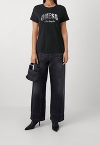 Kvinde iført sort "Guess Los Angeles" t-shirt, brede mørke jeans, sorte spidse støvler, holder en lille sort håndtaske.