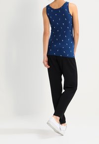 Persona vestida con una blusa sin mangas de color azul marino con pequeños estampados blancos, pantalones negros y zapatillas blancas, de pie de espaldas al espectador.