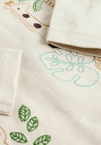 Lichtbeige stof met verschillende kleurrijke borduursels van een giraffe, groene bladeren en abstracte vormen in blauw en bruin. Zachte textuur.