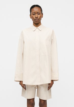 Frau trägt beige-weiß gestreiftes, übergroßes Hemd mit passenden knielangen Shorts vor einem einfarbigen hellen Hintergrund.