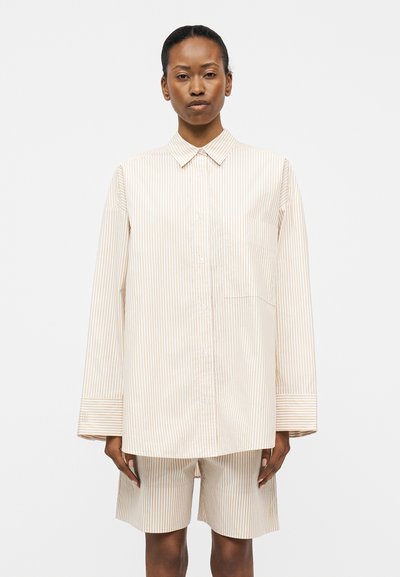 Femme portant une chemise oversize beige et blanche à rayures verticales avec un short assorti jusqu’aux genoux sur un fond clair uni.