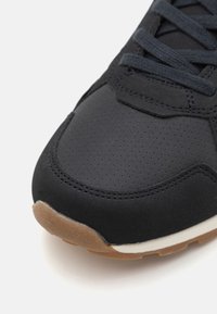 Black athletic shoe with perforated upper, textured material, and dark laces. Features a contrasting gum rubber outsole and white midsole.