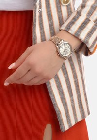 Reloj de pulsera plateado con una cara redonda, correa de metal y características de cronógrafo. Combinado con una chaqueta a rayas y una falda roja acanalada.