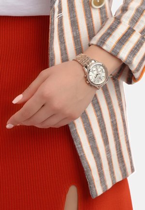 Orologio da polso argentato con quadrante rotondo, bracciale in metallo e funzioni di cronografo. Abbinato a un blazer a righe e una gonna rossa a coste.