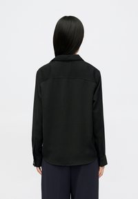 Žena s ravnom crnom kosom, obučena u crnu majicu s dugim rukavima i tamne hlače, prikazana sa stražnje strane na jednostavnoj bijeloj pozadini.