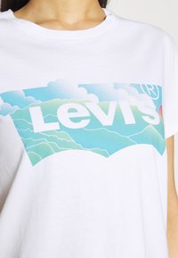 Vit bomull t-shirt med en stor, färgglad Levi's logotypgrafik med blå himmel och gröna kullar. Korta ärmar, rund halsringning.
