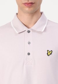 Polo shirt violet clair en tissu texturé, avec un col rayé blanc, trois boutons gris et un logo brodé jaune sur la poitrine.