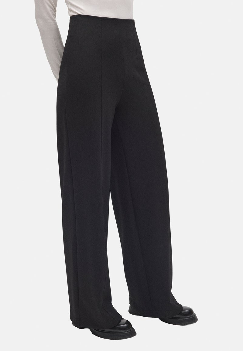 Mujer vestida con pantalones negros de pierna ancha y cintura alta, combinados con zapatos negros y una camiseta de manga larga gris claro, de pie contra un fondo blanco.