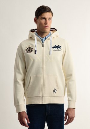 Sudadera cremosa con cremallera y capucha, bolsillos frontales y logotipos bordados. Presenta rayas en contraste en el interior de la capucha y puños acanalados.