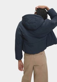 Marineblå pufferjakke med hætte, der har quiltet tekstur og elastiske manchetter, båret over beige vide bukser.