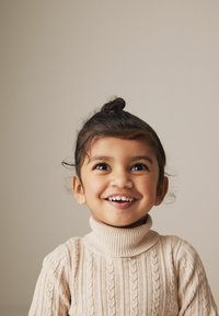 Besidintis vaikas su šypsena, vilkintis šviesiai rudą, švelnų turtleneck megztinį su tekstūruotu pynimu. Plaukai rišti kuoduku, su laisvais sruogomis.