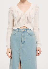 Beżowy, dziergany, krótki kardigan z dekoltem w serek, z przodu zapinany na guziki, z prążkowanymi mankietami, noszony na jasnoniebieską spódnicę dżinsową z bocznym rozcięciem.