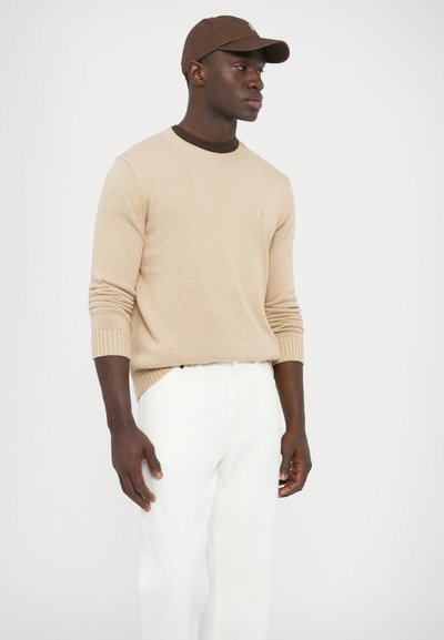 Béžový pletený sveter s okrúhlym výstrihom, žíhanými manžetami a malým logom, skombinovaný s bielymi nohavicami a hnedou čiapkou.