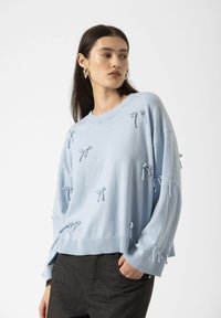 A woman with long dark hair wears a light blue sweater adorned with bow details, paired with dark pants, against a neutral background.
