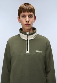 Olive green fleece pullover with a high neckline and cream-colored zipper. Features a small logo on the chest and textured fabric.