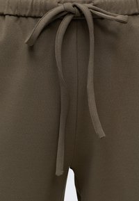 Gros plan d’un pantalon vert olive avec un cordon en tissu noué à la taille élastique montrant la texture et les détails des coutures.