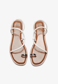 Hvide flade sandaler med flettede stropper og en skinnende sølvspænde på en gennemsigtig sål med en tan fodseng. Glat tekstur hele vejen igennem.