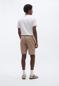 Καφέ λινά σορτς με τσέπη στο πίσω μέρος, συνδυασμένα με μονόχρωμη λευκή μπλούζα και πράσινα αθλητικά παπούτσια με λευκές και μωβ λεπτομέρειες.