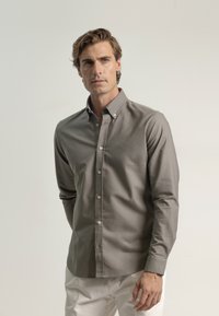 Camisa de botones gris de manga larga con cuello puntiagudo, que presenta botones blancos y un logo sutil en el pecho. Material suave y ligero.