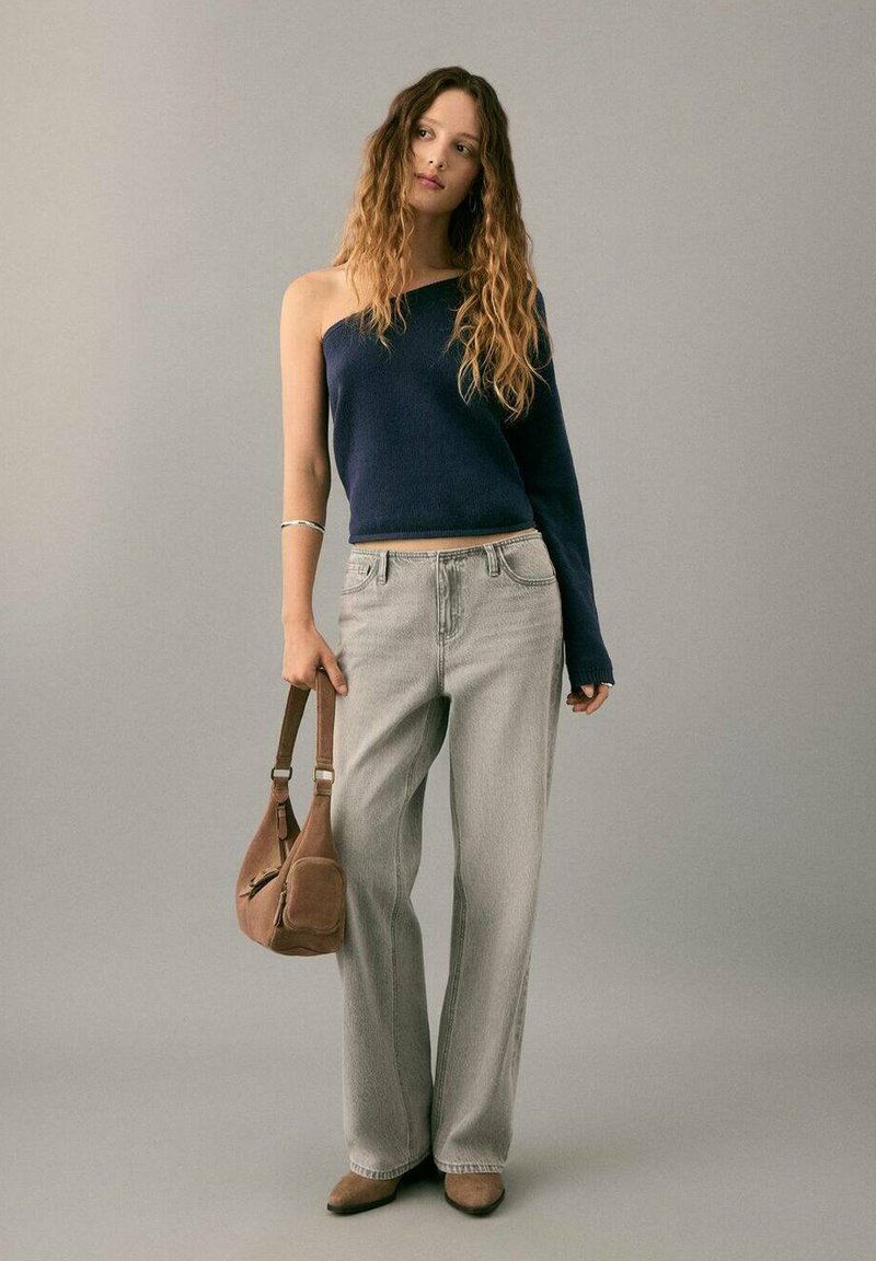 Marineblauer asymmetrischer Pullover, hellgraue Jeans mit weitem Bein, braune Schultertasche und beigefarbene Schuhe. Der Pullover hat eine strukturierte Oberfläche und eine lockere Passform.