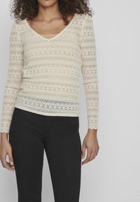 Kremfarget blonde topp med lange ermer, dyp V-hals og blomstermønster. Brukt over en beige singlet, sammen med svarte bukser. Strukturell, delikat design.