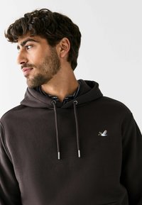 Brązowa bluza z kapturem i sznurkami, z małym haftowanym wzorem ptaka na piersi. Miękka tekstura i luźny krój.