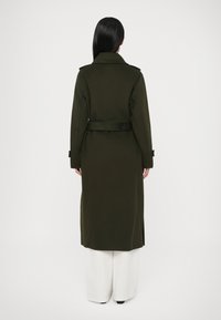 Lange olijfgroene jas met kraag, rugceintuur en zijsplitsingen. Gladde textuur met een getailleerde pasvorm, gecombineerd met witte brede broekspijpen.