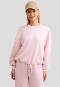 Sudadera rosa con cuello redondo, hombros caídos, cintura elástica y cordón ajustable. Tejido suave con una textura lisa.