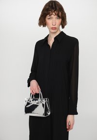Bolso de acrílico transparente con doble asa, forma rectangular y acabado brillante, sostenido en una mano, combinado con una camiseta negra de manga larga.