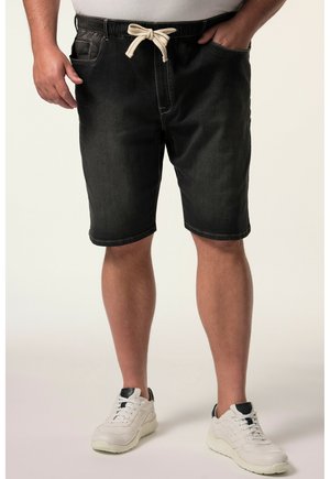 Uomo che indossa pantaloncini neri in denim fino al ginocchio con cordoncino bianco, t-shirt bianca e sneakers bianche su sfondo semplice.