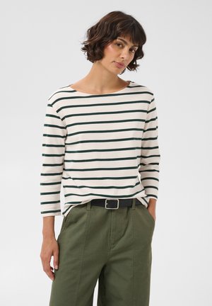 Femme aux cheveux courts et foncés portant une chemise blanche à manches longues avec des rayures horizontales noires et un pantalon vert avec une ceinture noire.