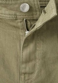 Primo piano di pantaloni in tessuto kaki con una cerniera argentata parzialmente aperta e un bottone argentato inciso con del testo.