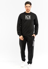 Felpa di cotone nera con logo "ICE" e pantaloni jogger neri abbinati, dotati di tasche laterali e grafiche bianche. Modello con sneakers bianche.