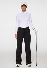 Vit långärmad idrottströja och svarta byxor, med en tight passform. Tillbehör inkluderar en vit keps och en golfklubba i handen.