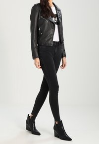 Chaqueta de motorista de cuero negra con cremalleras plateadas, camiseta gráfica blanca debajo, jeans ajustados negros y botas negras con cuña.