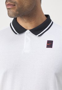 Polo bianco con un motivo a rete testurizzata, colletto nero con strisce bianche, placket a due bottoni e un logo quadrato Nike in rosso e nero.