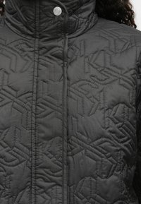 Čierna prešívaná bunda s vysokým golierom, s geometrickým vzorom a zapínaním na cvoky. Textúra vyzerá hladko a lesklo.