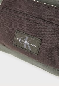 Tmavohnedý textilný vak s zeleným dnom. Obsahuje obdĺžnikový nášivku s nápisom "Calvin Klein Jeans" a strieborné logo "CK". Uzatváranie na zips.