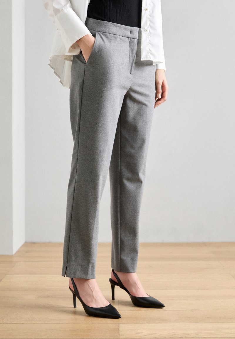 Femme portant un pantalon gris sur mesure, un top noir sans manches, une chemise blanche et des chaussures à talons aiguilles noires, debout sur un sol en bois.