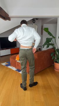 Άνδρας που στέκεται σε σαλόνι και τραβάει ένα λευκό φούτερ πάνω από το κεφάλι του, φορώντας ελαι πράσινο παντελόνι και μαύρα παπούτσια.