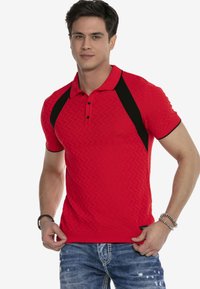 Rotes gestricktes Polohemd mit einem strukturierten Zickzackmuster, schwarzen Akzenten an den Schultern und einer Drei-Knopf-Leiste. Getragen mit blauen Jeans.