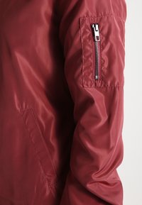 Červená bomber bunda s hladkou, lesklou texturou. Vybavena kapsou na zip na rukávu a boční kapsou, která zdůrazňuje funkční designové prvky.