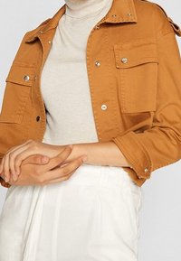 Brun denimjakke med sølvfarvede trykknapper, to brystlommer og en afslappet pasform. Båret over en lys beige turtleneck.